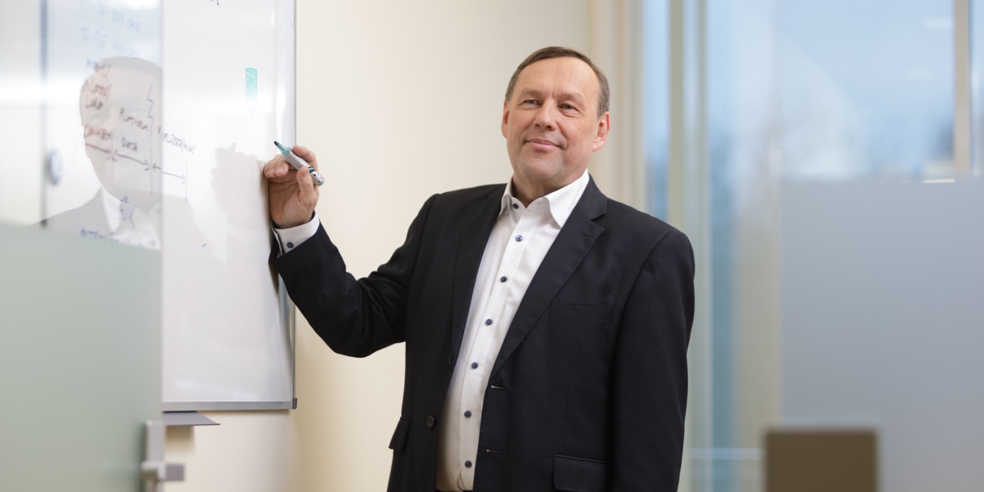 Person in a dark business suit stands beside a whiteboard, holding a marker and pointing toward a hand-drawn diagram during a presentation or planning session in a modern office. Large windows and glass partitions are visible in the background.